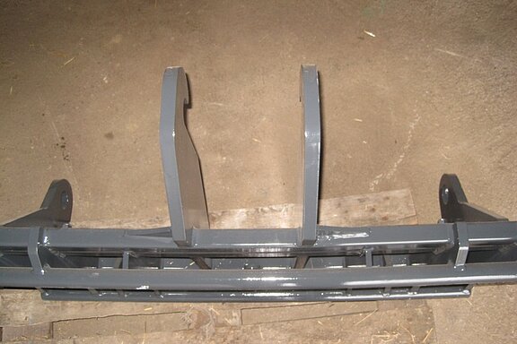 Graue Metallhalterungskonstruktion mit zwei senkrechten Stützen, die auf einer Holzpalette auf einem Betonboden steht.