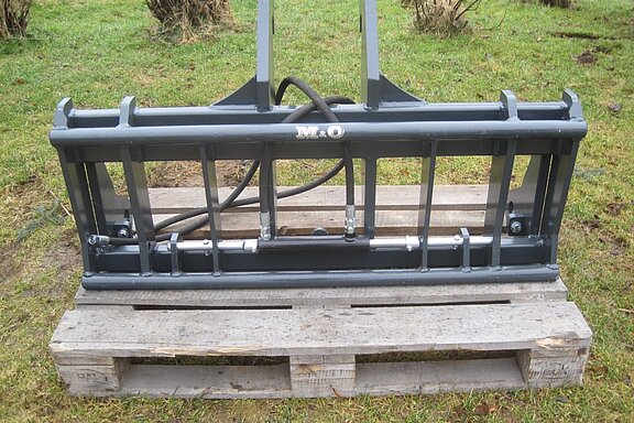 Hydraulik-Anbaugerät aus Metall auf einer Holzpalette, platziert auf einer Rasenfläche mit Sträuchern im Hintergrund.